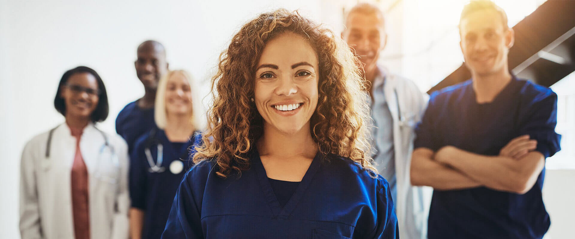 The image shows a group of healthcare professionals posing together with a smiling woman in the foreground, who appears to be a nurse, standing out from the others due to her direct gaze at the camera.