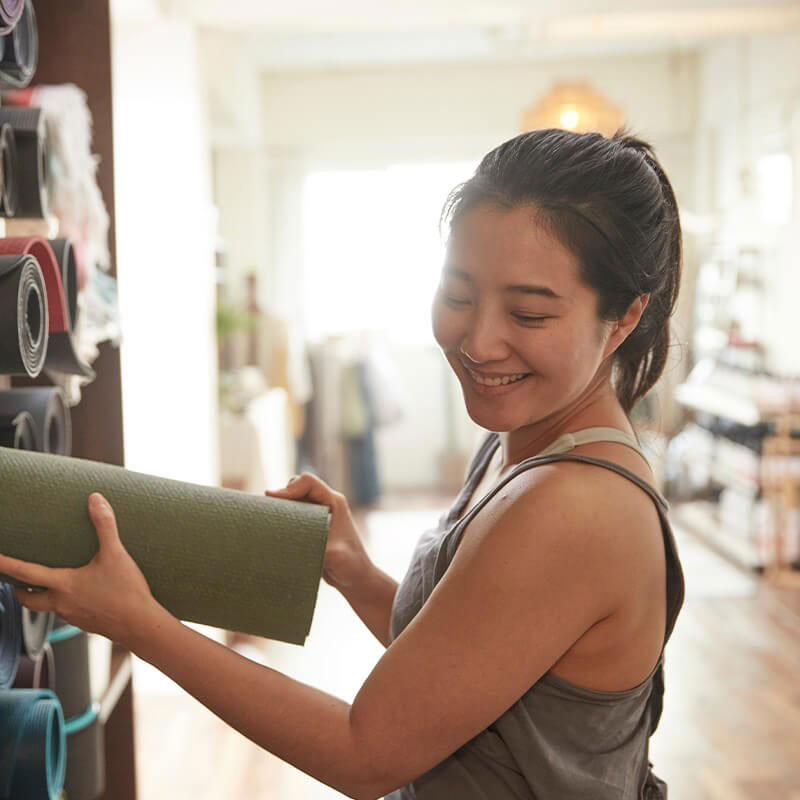 A woman in a yoga studio holding a rolled-up yoga mat with a smile on her face.