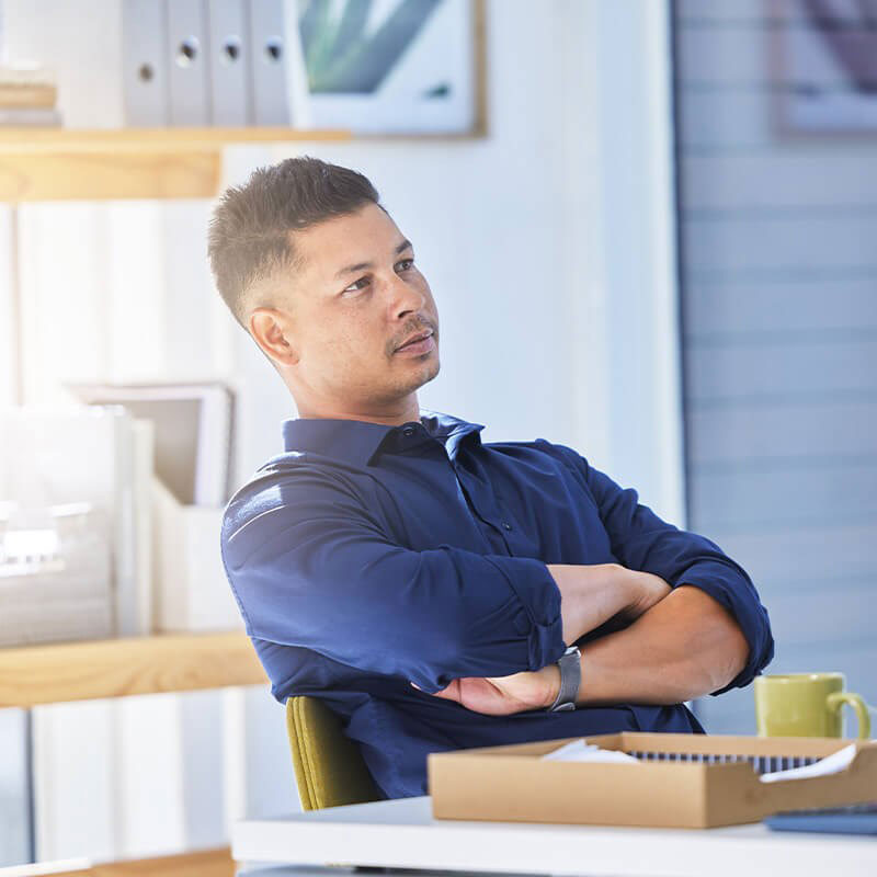 The image shows a man sitting at a desk with his arms crossed, looking off to the side, with a relaxed posture in an office environment.