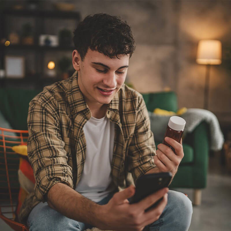 A young man sitting on a couch, smiling at his smartphone while holding a bottle of pills, with a plaid shirt and casual attire.