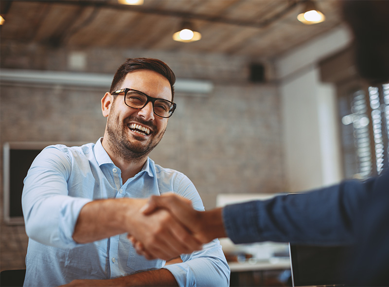 The image shows two individuals shaking hands over a desk in an office environment.
