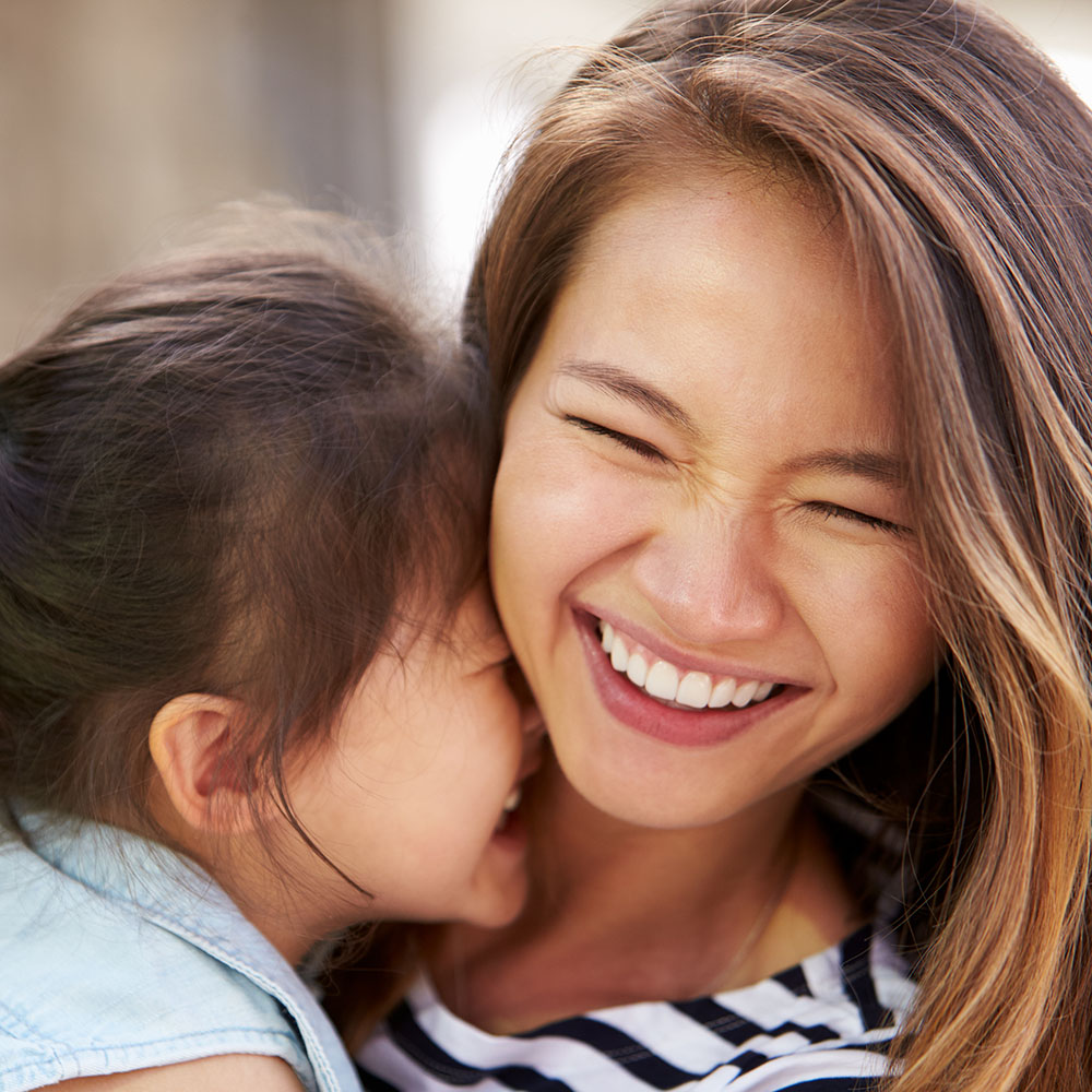 A woman with long hair, smiling broadly, hugs a child who is also smiling at her.